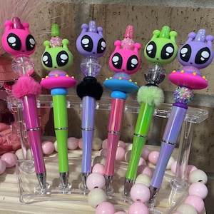 May include: Six colorful pens with alien and spaceship themed toppers. The pens have iridescent barrels and are displayed on a white stand with pink and white beads.
