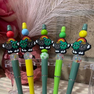 May include: Five green and yellow pens with a black leprechaun charm on top. The charms have a pot of gold, a rainbow, and a four-leaf clover.