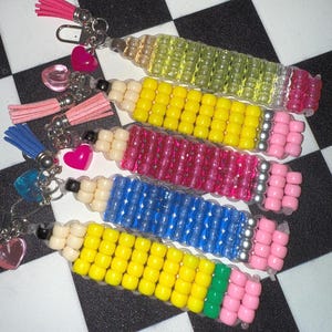 May include: Four beaded pencil keychains in yellow, pink, blue, and green. Each keychain has a different color of beads for the pencil body, a beige eraser, and a pink eraser tip. Each keychain has a tassel and heart charm.