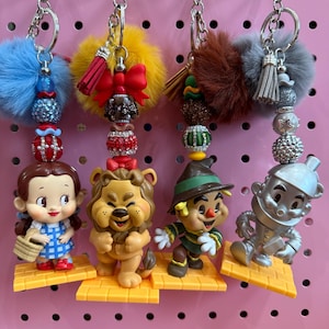 May include: Keychains featuring characters from The Wizard of Oz. Each keychain has a character figurine, decorative beads, and a fluffy pom-pom. Characters include Dorothy, the Cowardly Lion, the Scarecrow, and the Tin Man. The keychains hang on a pink pegboard.