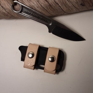 May include: A black drop forged steel knife with a black blade and a tan leather sheath. The knife has a ring at the end of the handle. The sheath has two tan leather straps with silver snaps.