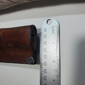 May include: A brown leather sheath with a metal snap closure. The sheath is measured with a ruler, showing it is 6.35 cm long.