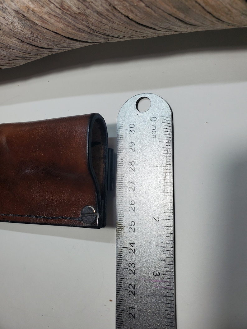 May include: A brown leather sheath with a metal snap closure. The sheath is measured with a ruler, showing it is 6.35 cm long.