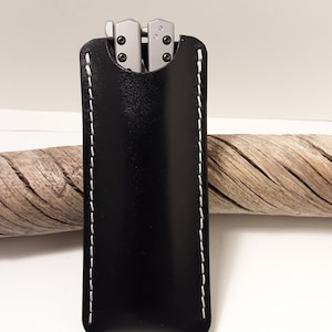 May include: A black leather knife sheath with white stitching. The sheath has a flap that covers the top of the knife.