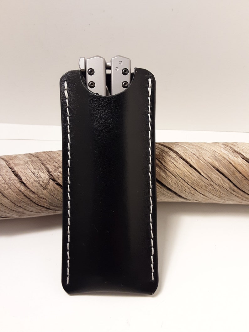 May include: A black leather knife sheath with white stitching. The sheath has a flap that covers the top of the knife.