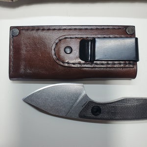 May include: A brown leather sheath with a black metal clip and a silver knife with a black carbon fibre handle. The knife has a small, pointed blade and the text "P15002A" is visible on the handle.