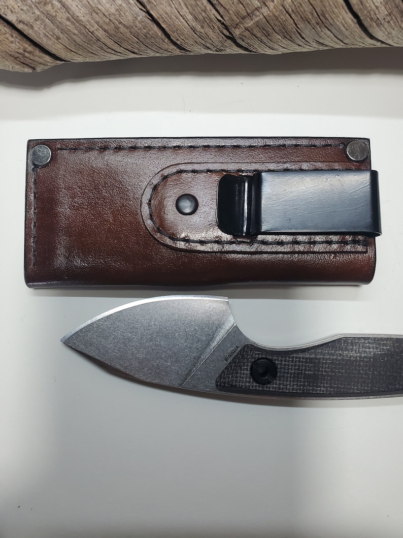 May include: A brown leather sheath with a black metal clip and a silver knife with a black carbon fibre handle. The knife has a small, pointed blade and the text "P15002A" is visible on the handle.