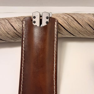 May include: A brown leather knife sheath with white stitching. The sheath has a flap that folds over to secure the knife inside.