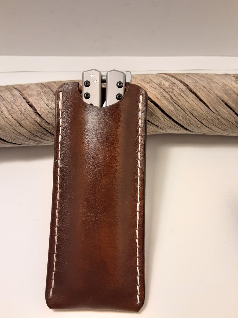May include: A brown leather knife sheath with white stitching. The sheath has a flap that folds over to secure the knife inside.