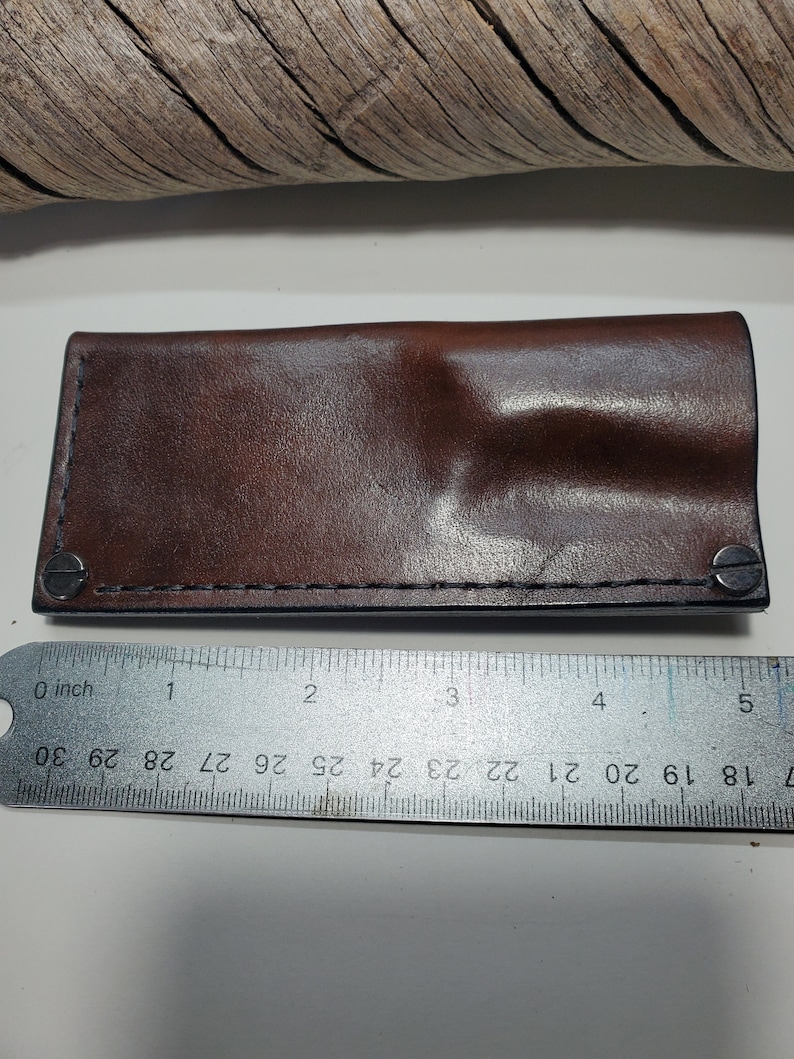 May include: A brown leather knife sheath with a stitched seam and two metal rivets. The sheath is measured with a ruler in inches.