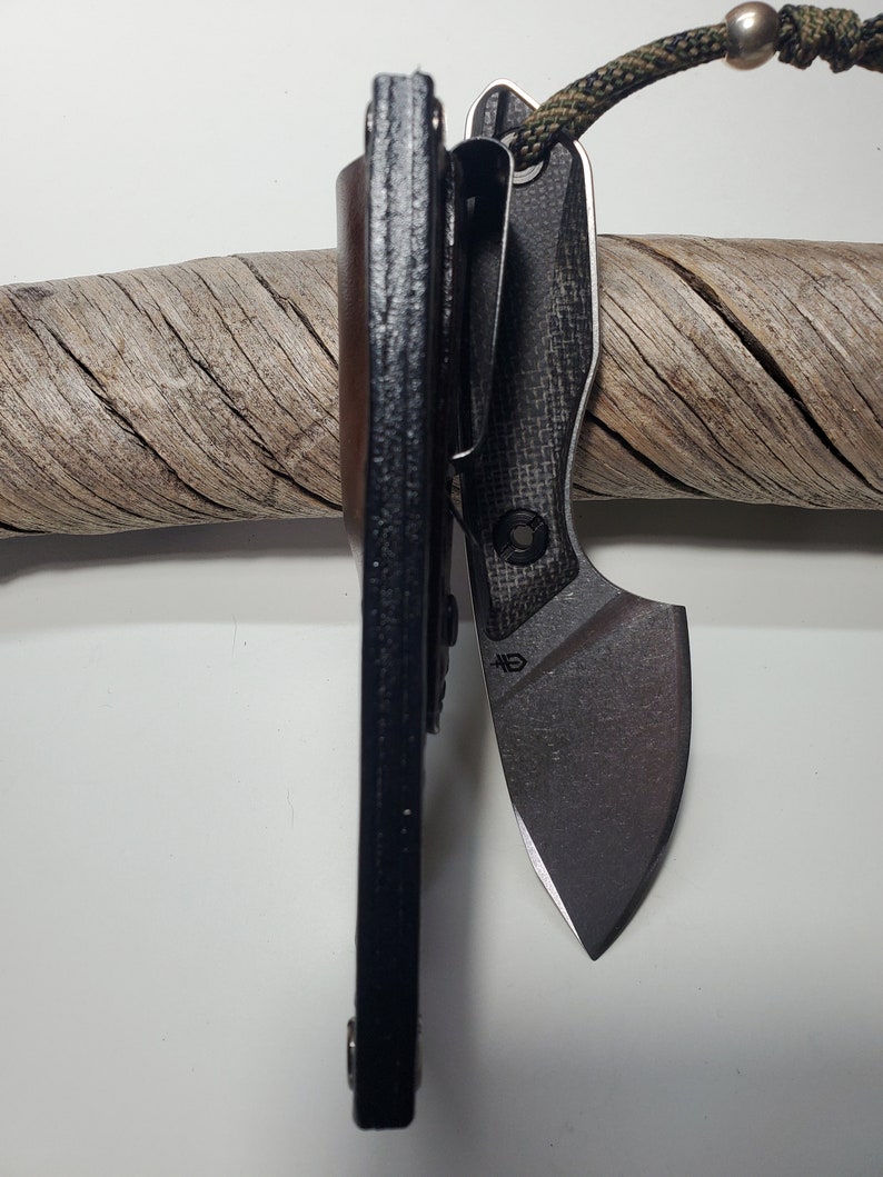 May include: A black leather knife sheath with a carbon fibre pocket knife with a black blade. The knife is attached to a green and brown lanyard.