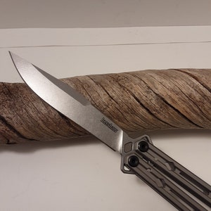 May include: A silver Kershaw brand butterfly knife with a partially open blade. The blade is sharp and has a slight curve. The handle is made of metal and has a textured design.