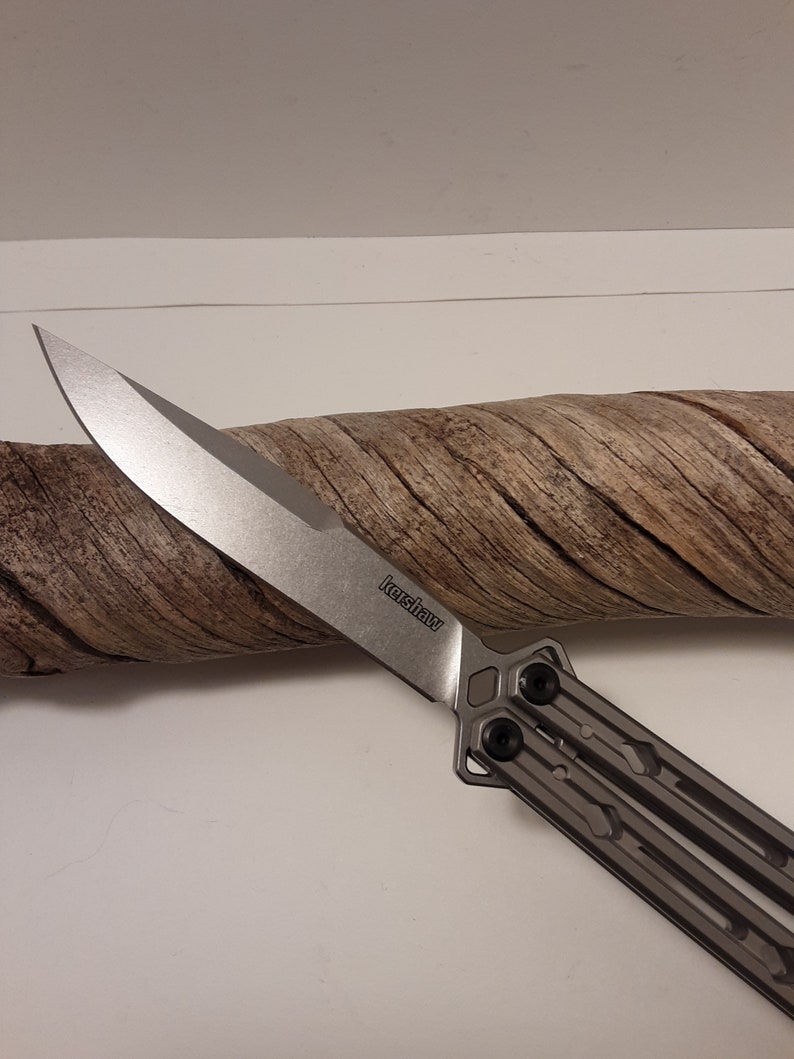 May include: A silver Kershaw brand butterfly knife with a partially open blade. The blade is sharp and has a slight curve. The handle is made of metal and has a textured design.