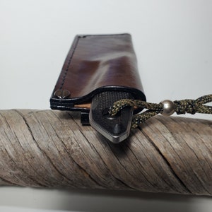 May include: A brown leather sheath with black stitching detail and a silver rivet. A black and silver knife is inside the sheath with a green and black cord attached.