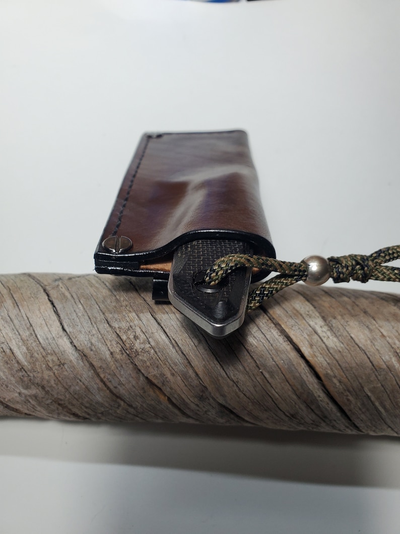 May include: A brown leather sheath with black stitching detail and a silver rivet. A black and silver knife is inside the sheath with a green and black cord attached.