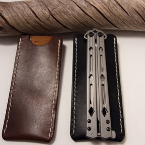 May include: Two leather sheaths for a butterfly knife. The sheath on the left is brown with white stitching. The sheath on the right is black with white stitching. A silver butterfly knife is inside the black sheath.