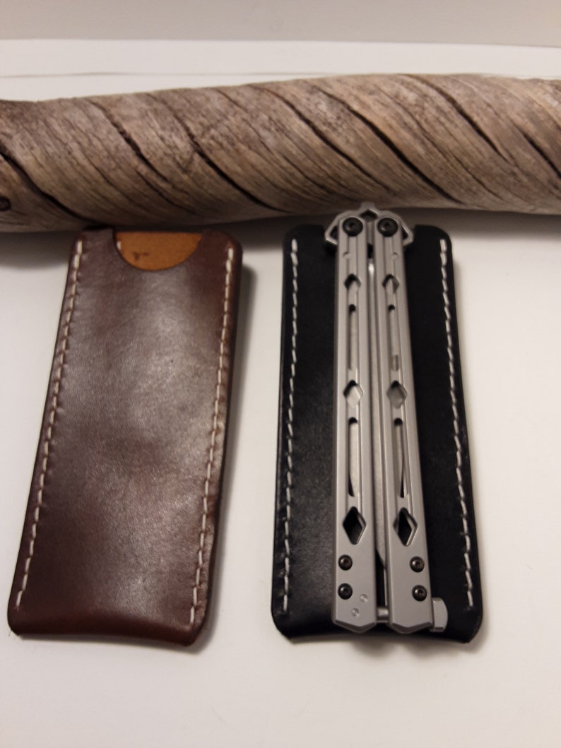 May include: Two leather sheaths for a butterfly knife. The sheath on the left is brown with white stitching. The sheath on the right is black with white stitching. A silver butterfly knife is inside the black sheath.