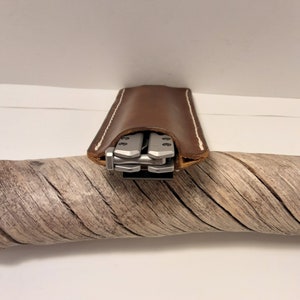 May include: A brown leather case with white stitching, designed to hold a multi-tool. The multi-tool is visible inside the case.