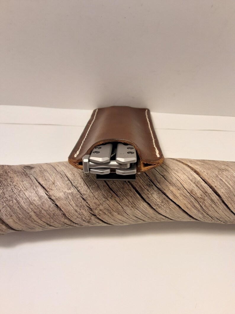 May include: A brown leather case with white stitching, designed to hold a multi-tool. The multi-tool is visible inside the case.