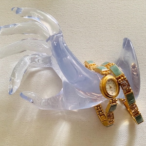 May include: A gold-toned wristwatch with a white face and gold hands, featuring a jade-colored band. The watch is displayed on a clear, hand-shaped display stand. The watch has a round face and a delicate design.