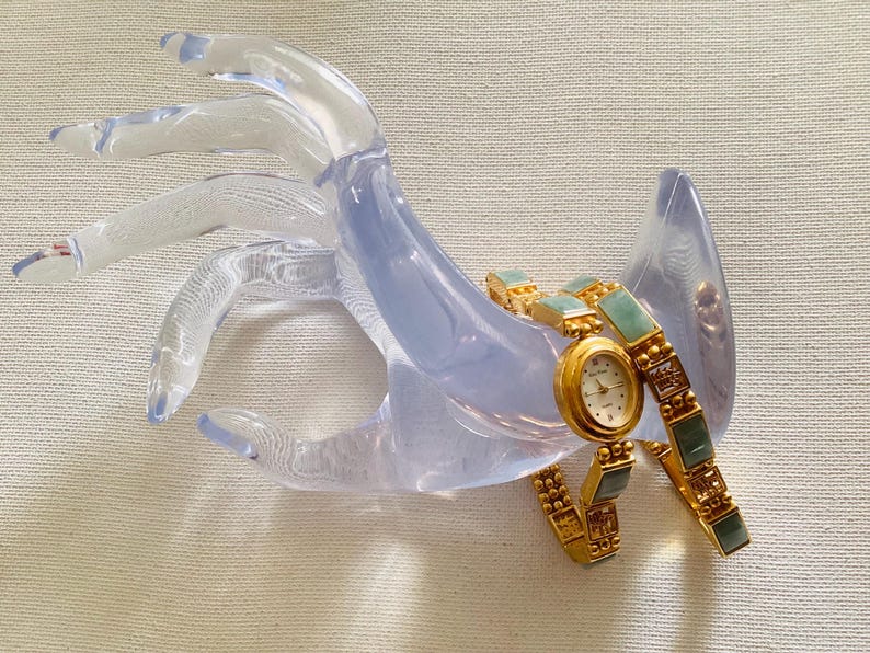 May include: A gold-toned wristwatch with a white face and gold hands, featuring a jade-colored band. The watch is displayed on a clear, hand-shaped display stand. The watch has a round face and a delicate design.