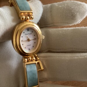 May include: A gold-toned quartz watch with an oval face and a light-colored dial. The watch features Roman numerals and the brand name "Gen Time". The watch band has rectangular, light-green jade-like segments.