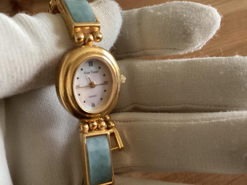 May include: A gold-toned quartz watch with an oval face and a light-colored dial. The watch features Roman numerals and the brand name "Gen Time". The watch band has rectangular, light-green jade-like segments.