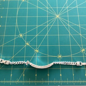 K&ouml;nnte beinhalten: Silberfarbenes Armband mit einem Panzerketten-Design und einem dekorativen Mittelteil. Das Armband liegt auf einer gr&uuml;nen Schneidematte mit Zentimetermarkierungen.