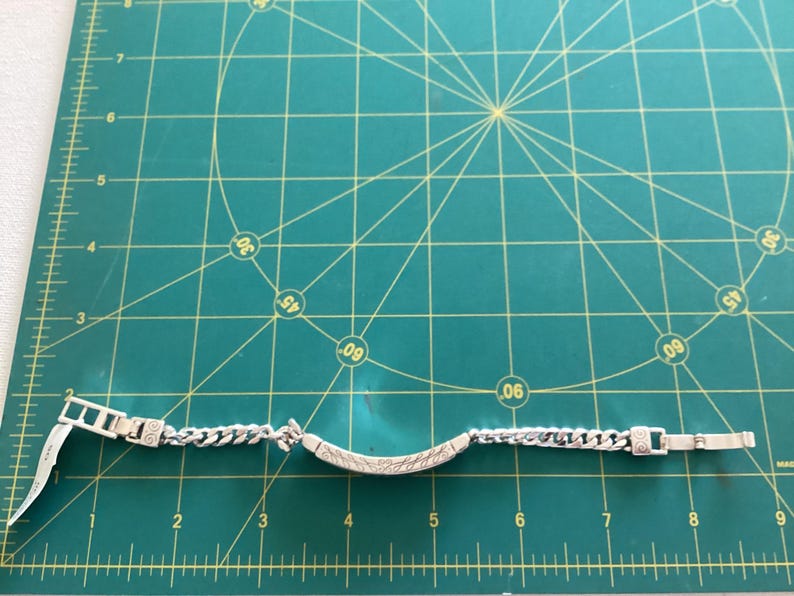 K&ouml;nnte beinhalten: Silberfarbenes Armband mit einem Panzerketten-Design und einem dekorativen Mittelteil. Das Armband liegt auf einer gr&uuml;nen Schneidematte mit Zentimetermarkierungen.