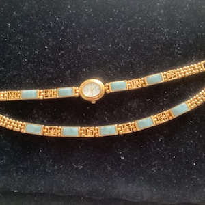 May include: Gold-toned wristwatch with a light blue rectangular band and a small oval face. The watch band is separated into two pieces, displaying the intricate design and the watch's delicate features.