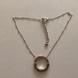 May include: A silver necklace with a round pendant featuring a red and white design. The pendant is a circle with a small opening in the center.