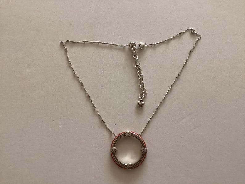 May include: A silver necklace with a round pendant featuring a red and white design. The pendant is a circle with a small opening in the center.