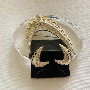 May include: A vintage jewelry set featuring a gold-toned brooch and matching earrings. The brooch has a curved design with clear rhinestones and a scalloped edge. The earrings are crescent-shaped with similar detailing.