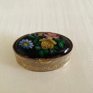 May include: An oval-shaped metal trinket box featuring a black lid decorated with a painted floral motif of yellow, pink, and blue flowers with green leaves. The sides of the box are finished in a silver tone with an embossed decorative pattern.