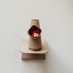 May include: A gold-toned ring with a large, faceted, red-orange gemstone. The gemstone is set in a twisted rope-like frame. The ring is displayed on a beige ring holder.