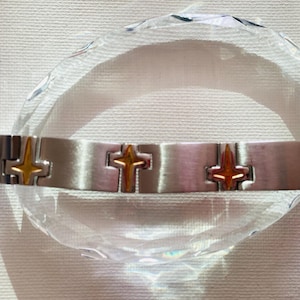 Puede incluir: Un brazalete plateado con detalles de cruces doradas. El brazalete, compuesto por eslabones rectangulares, se exhibe sobre un objeto transparente y facetado. Los detalles de las cruces añaden elegancia a la joya.