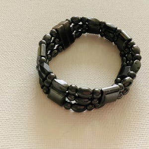 Puede incluir: Una pulsera gris oscuro con tres filas de cuentas y detalles rectangulares. La pulsera está hecha de pequeñas cuentas redondeadas y piezas geométricas más grandes. La pulsera está sobre una superficie beige claro.