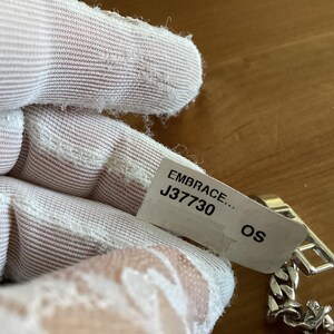 K&ouml;nnte beinhalten: Ein silbernes Kettenarmband mit rechteckigem Verschluss. Das Armband ist teilweise sichtbar, mit einem Etikett mit der Aufschrift "EMBRACE... J37730 OS". Das Armband liegt auf einer Holzoberfl&auml;che.