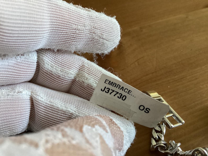 K&ouml;nnte beinhalten: Ein silbernes Kettenarmband mit rechteckigem Verschluss. Das Armband ist teilweise sichtbar, mit einem Etikett mit der Aufschrift "EMBRACE... J37730 OS". Das Armband liegt auf einer Holzoberfl&auml;che.