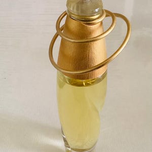 Puede incluir: Un frasco de perfume de vidrio con líquido amarillo pálido, que presenta una tapa cónica de color dorado texturizado adornada con dos anillos de alambre circulares y delgados de color dorado que rodean la parte superior, y un tapón transparente.