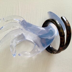 May include: A clear, plastic hand-shaped display stand holding two dark brown, tortoiseshell-patterned bangles. The stand is transparent, allowing the bangles to be the focal point. The bangles are approximately 2.5 inches in diameter.