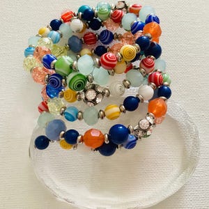May include: A multi-strand coil bracelet featuring a variety of round beads in shades of blue, orange, yellow, green, red, and white. The design includes translucent, opaque, and swirled glass beads interspersed with silver-toned metal spacers.