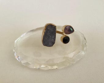 May include: A unique ring featuring a large, rough-cut, dark gray stone, a pear-shaped purple stone, and a small black stone. The stones are set on a gold-colored band. The ring is displayed on a clear, faceted, oval-shaped glass dish.