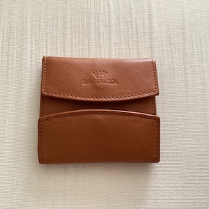 May include: A tan leather coin purse featuring a fold-over flap with a snap closure. The front of the flap is embossed with a small crown icon above the text DANIEL LODA. The wallet has a smooth texture and a rectangular shape with rounded corners.