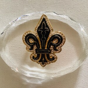 May include: A gold-toned fleur-de-lis brooch with black enamel detailing and encrusted with small, clear crystals. The brooch is set on a clear, oval-shaped glass or crystal base, resting on a white surface.