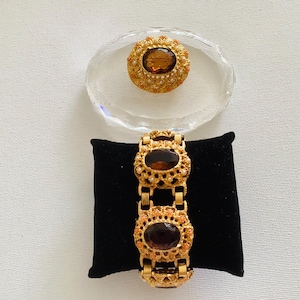Könnte beinhalten: Ein goldfarbenes Armband mit drei ovalen, dunkelbraunen Edelsteinen und eine passende Brosche mit einem ähnlichen Edelstein. Der Schmuck wird auf einer schwarzen Samtoberfläche und einer ovalen Glasschale präsentiert.