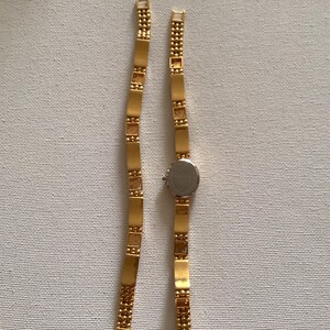 May include: A gold-toned wristwatch with a silver-colored face and a rectangular link band. The band is made of small, rectangular gold-colored links. The watch is displayed on a white surface.