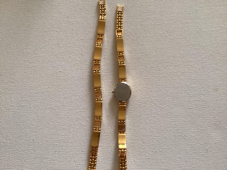 May include: A gold-toned wristwatch with a silver-colored face and a rectangular link band. The band is made of small, rectangular gold-colored links. The watch is displayed on a white surface.