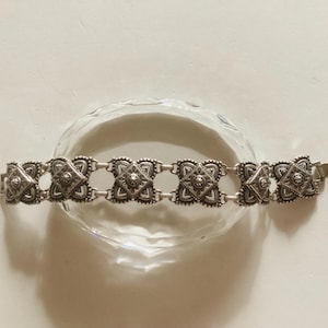May include: A silver bracelet with a decorative design featuring multiple square-shaped links. The bracelet has a clasp closure.