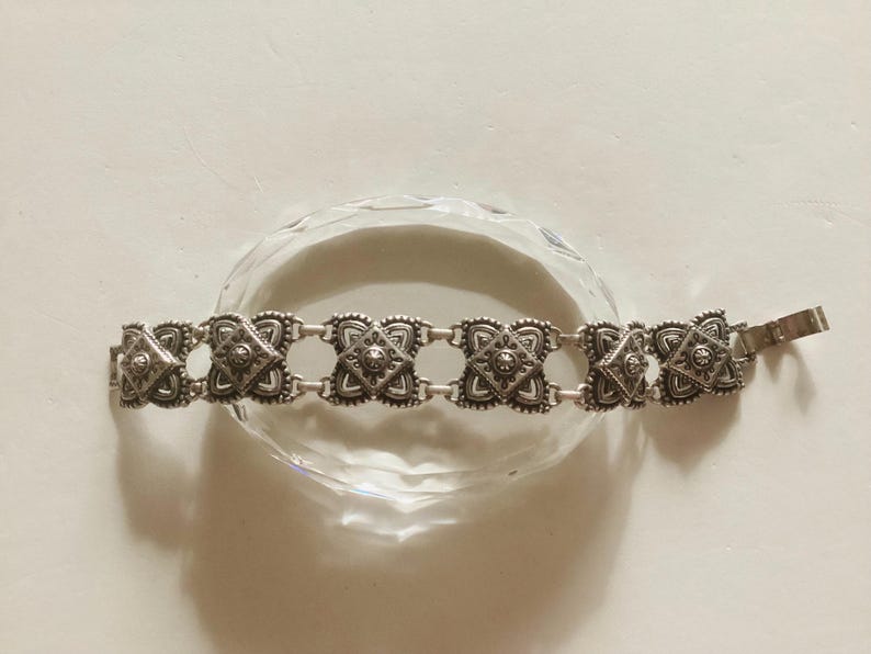 May include: A silver bracelet with a decorative design featuring multiple square-shaped links. The bracelet has a clasp closure.